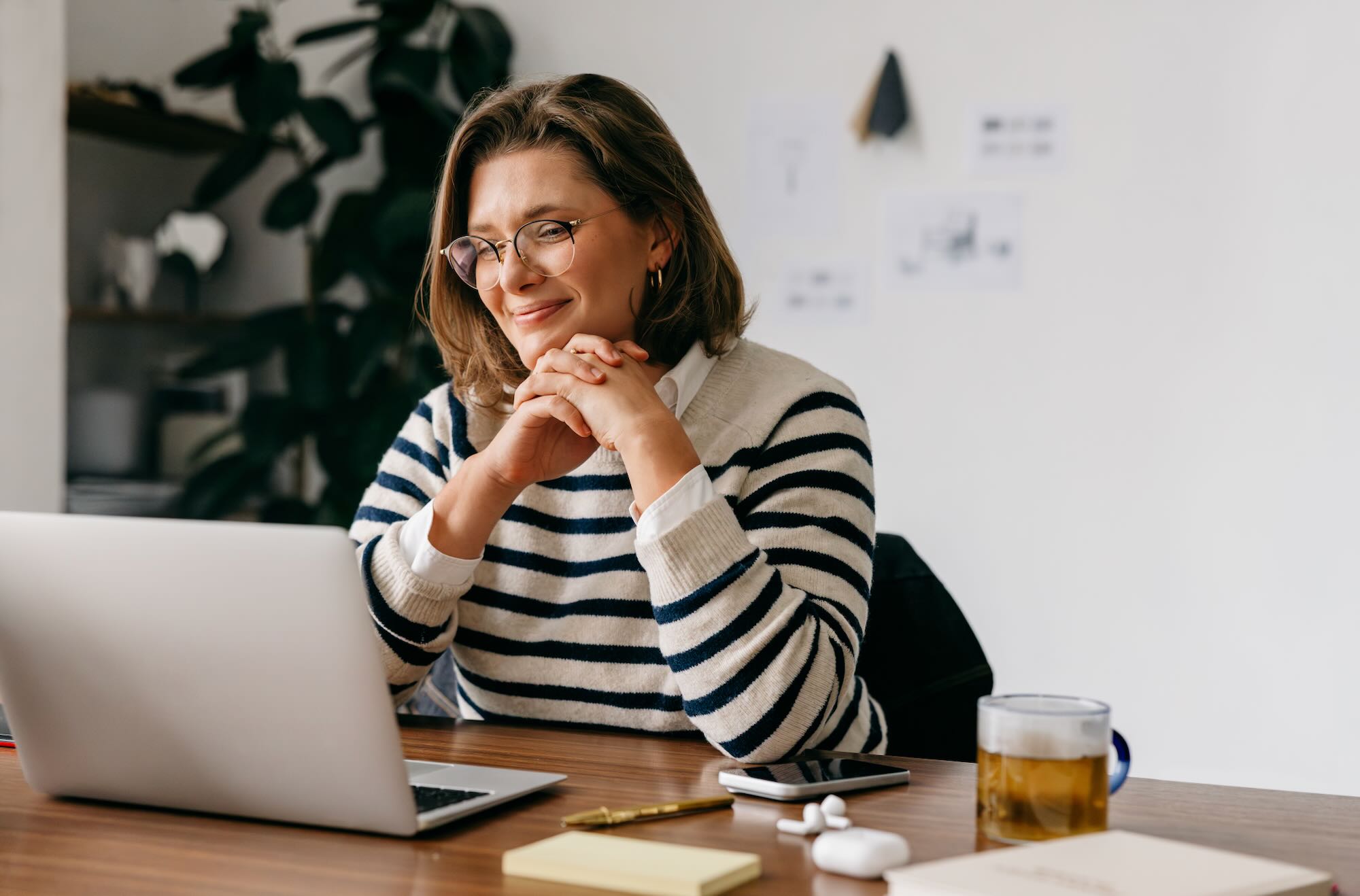 A smiling woman with glasses and a striped shirt sits at a desk with a laptop.